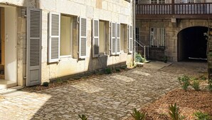 Une cour dallée de pierres, des bâtiments en pierre avec des fenêtres à volets et un balcon doté d’une balustrade en bois.