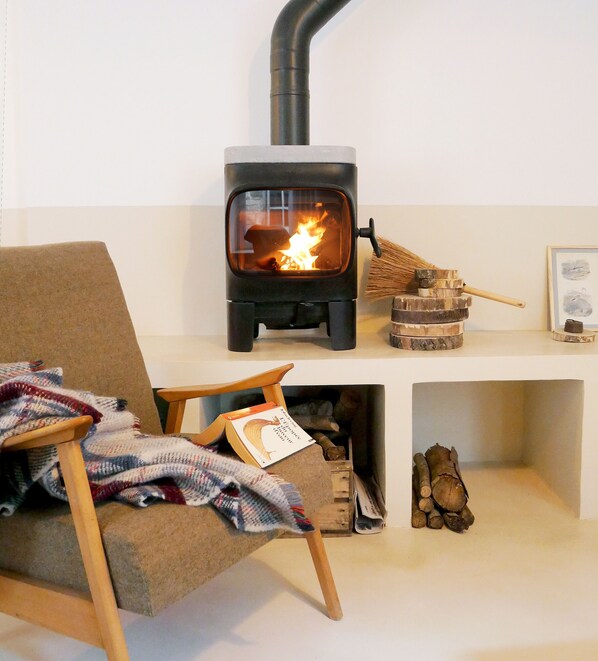A cozy room with a wood-burning stove, a chair with a blanket, and a bookshelf with books and decorative items. - Cozy N'est house on historic courtyard-garden, close to station and city center (Besançon)