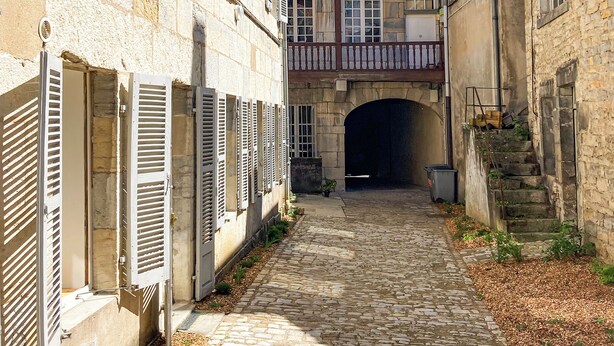 Une étroite ruelle pavée, bordée de bâtiments historiques en pierre aux fenêtres fermées par des volets et surmontée d’une arche centrale.