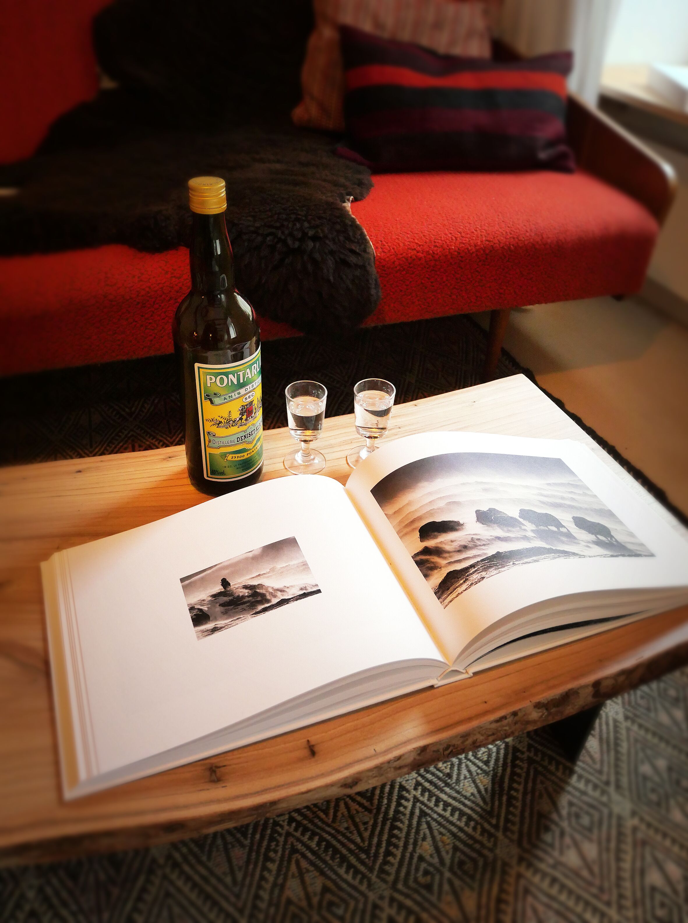 Una botella de Pontarj sobre una mesa de madera, con dos vasos para licor y un libro abierto con fotografías.