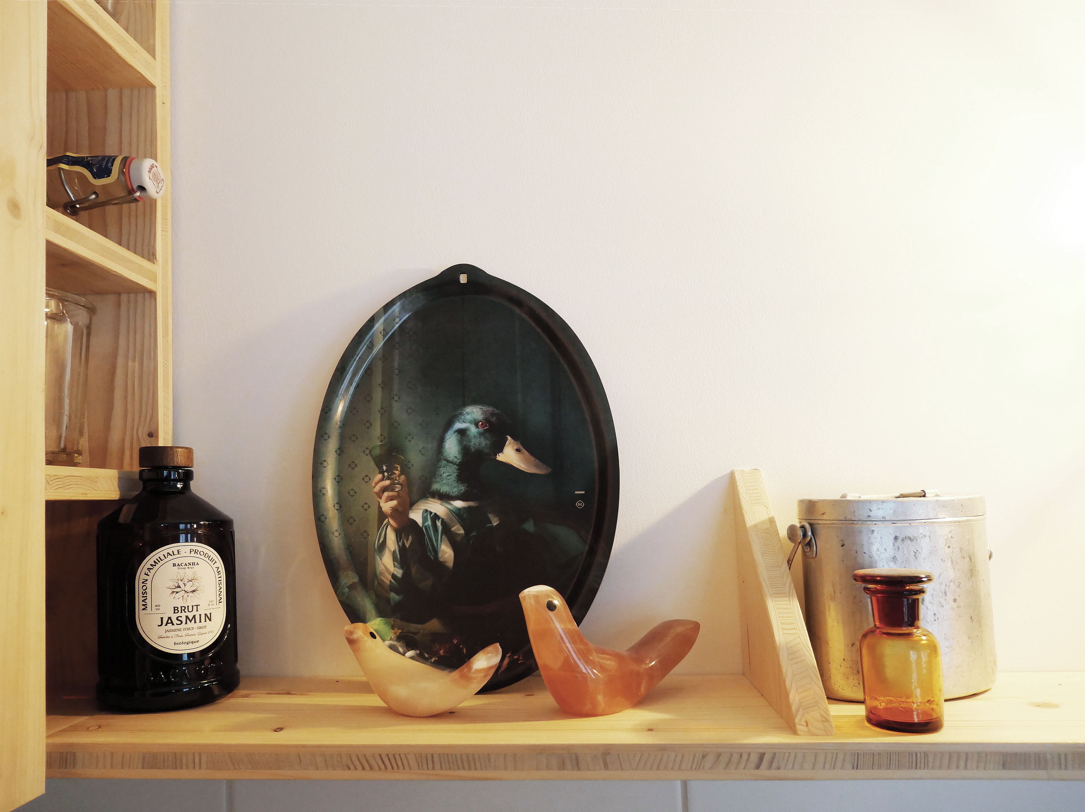 Uno scaffale in legno con una bottiglia di Brut Jasmin, un quadro a tema anatra e una piccola scultura di un uccellino arancione.