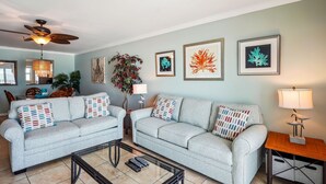 A living room with two grey sofas, a glass coffee table, a ceiling fan, and framed artwork on the wall. - Sea Breeze 106 - Sea, Sun, and Coastal Serenity (Madeira Beach)