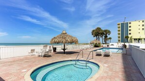 A pool area with a thatched umbrella, lounge chairs, and a view of the ocean. - Sea Breeze 106 - Sea, Sun, and Coastal Serenity (Madeira Beach)