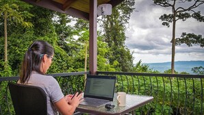 A person sitting at a table with a laptop and a smartphone, overlooking a lush green landscape with trees and a view of distant mountains. - Casa Bromelia, 30 minutes from La Fortuna (La Tigra)