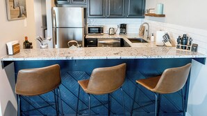 A modern kitchen with dark cabinets, a stainless steel refrigerator, a microwave, and a coffee maker. There are three bar stools at the island.
