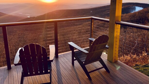 A wooden deck with two Adirondack chairs facing a scenic view of a sunset over a mountainous landscape.