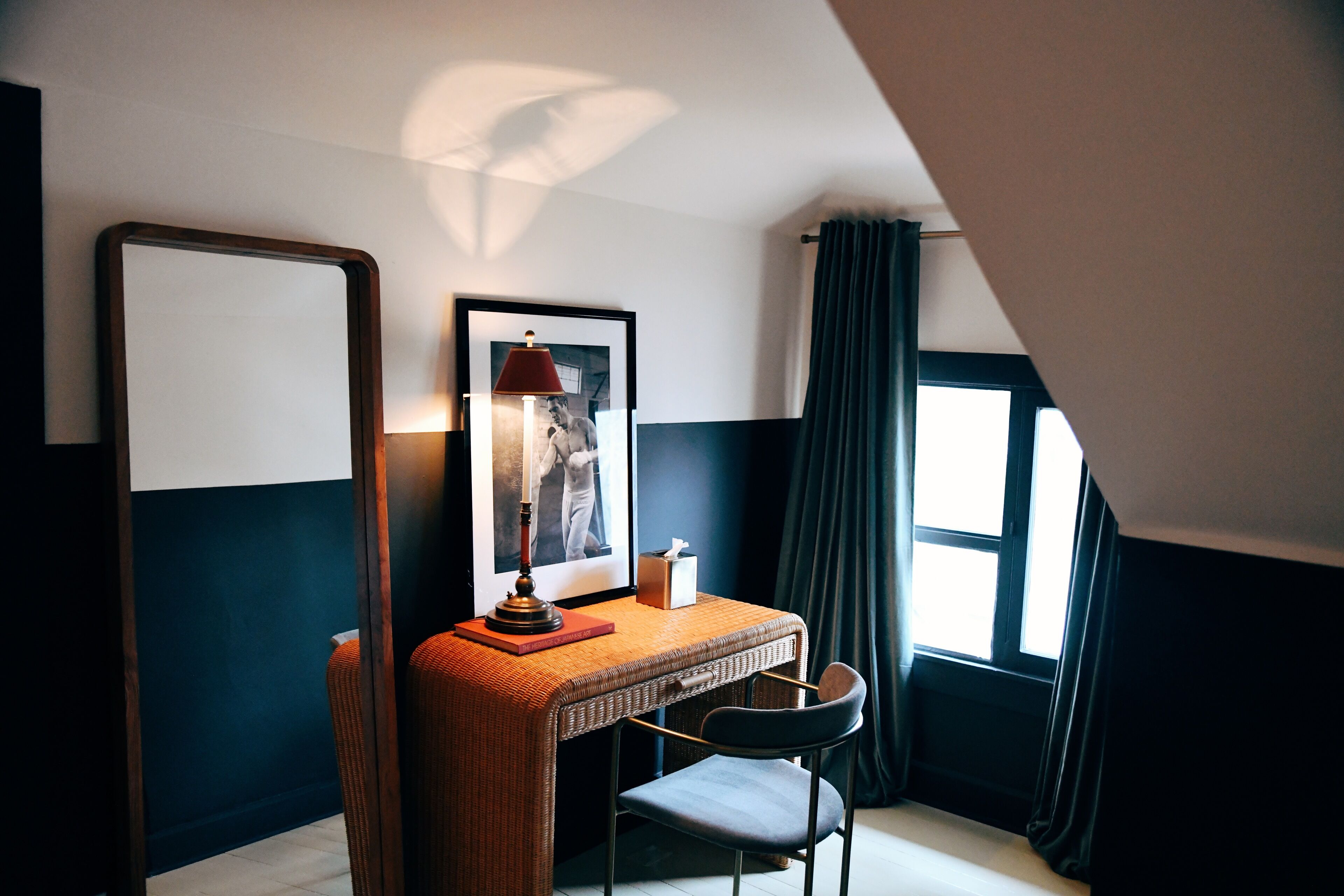 A room with a desk, chair, lamp, framed picture, and a window with curtains.