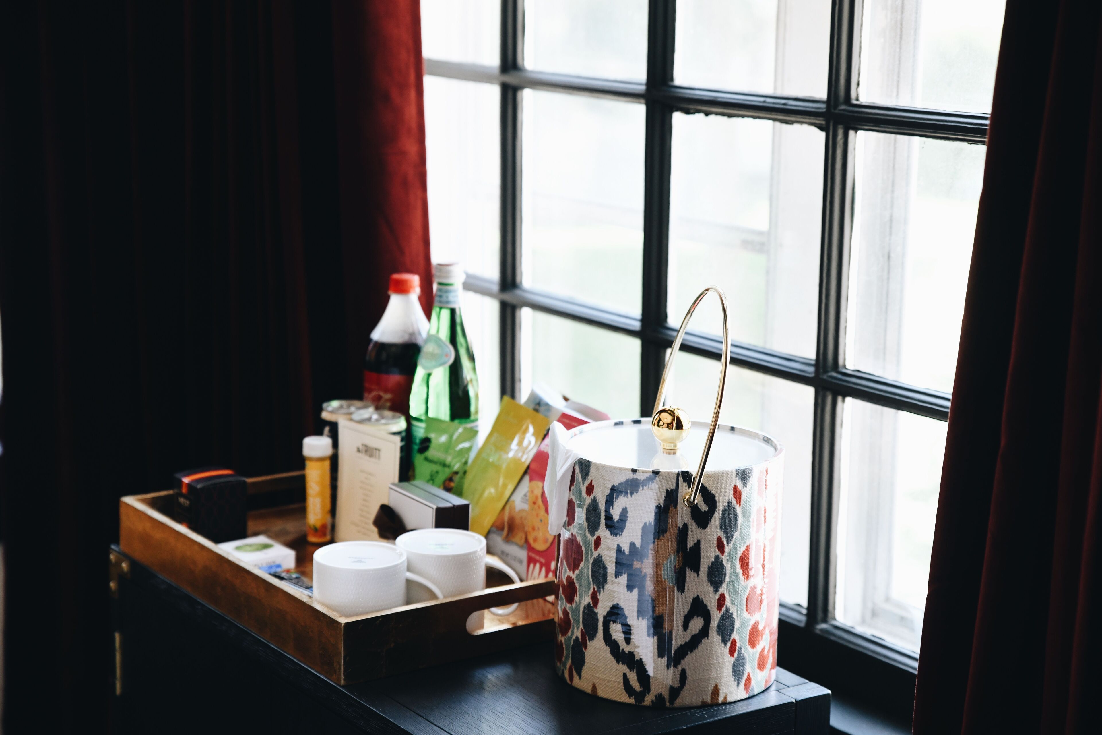 A tray with a patterned mug, a bottle, and some packaged snacks on a wooden table near a window with curtains.