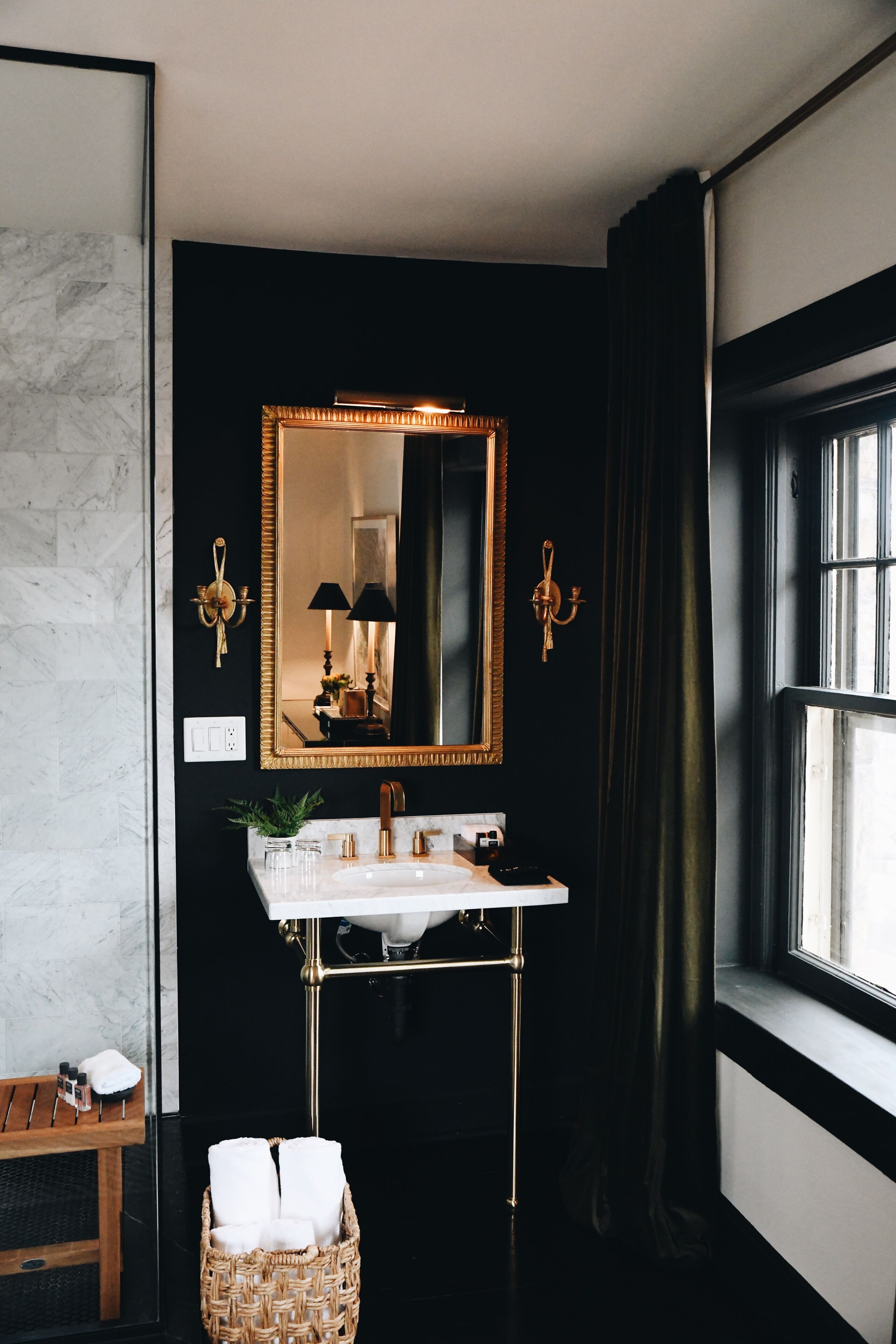 Een badkamer met een zwarte muur, een spiegel met gouden lijst, een wit wastafelmeubel en een raam met gordijnen.