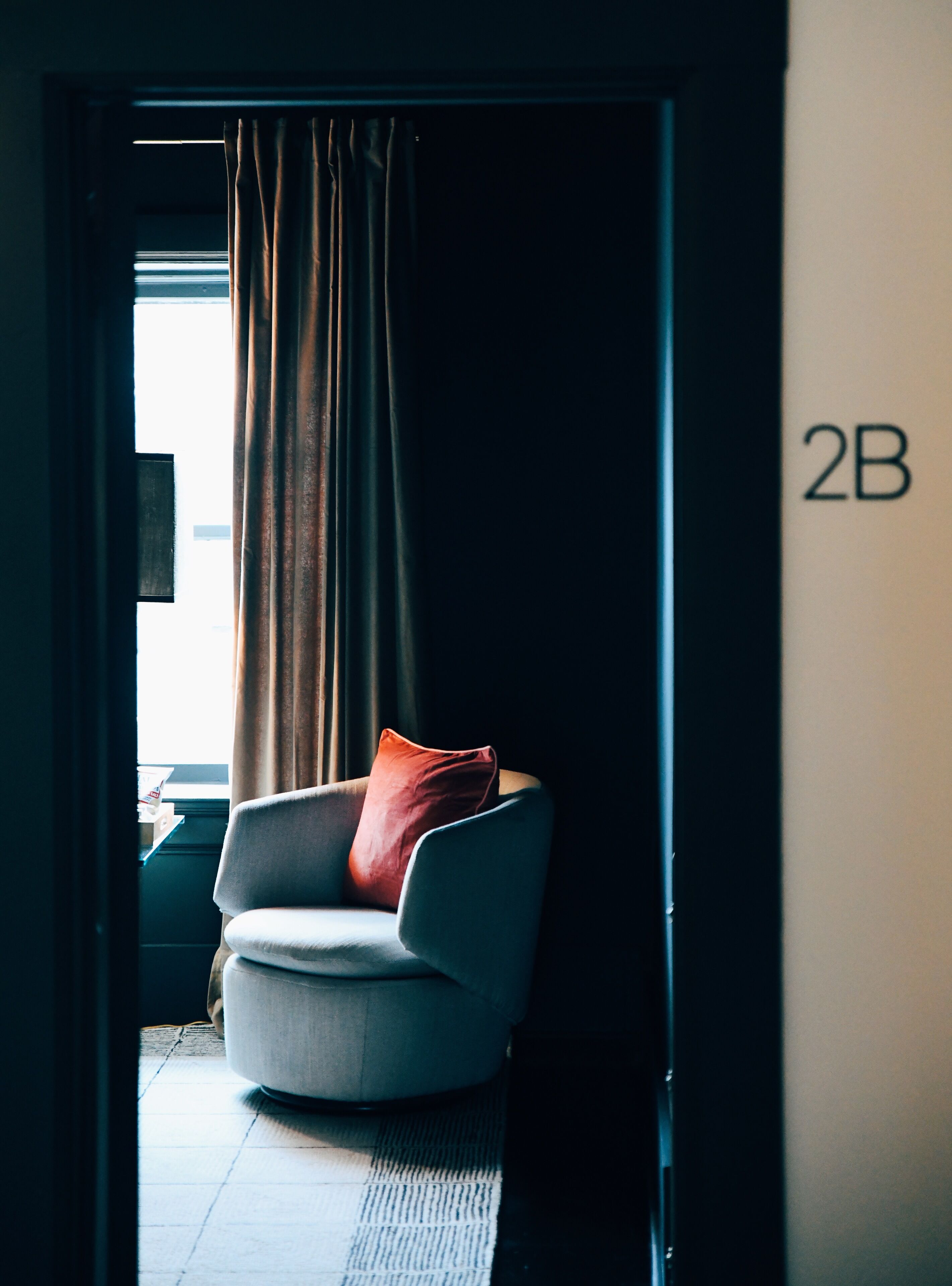 Ein modernes Zimmer mit einem grauen Sessel, einem roten Kissen und einem Fenster mit Vorhängen. Der Raum hat einen Fliesenboden und eine Tür mit der Aufschrift 2B.