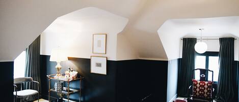 A room with a high ceiling, a chair, a small table with a lamp, and a window with curtains.