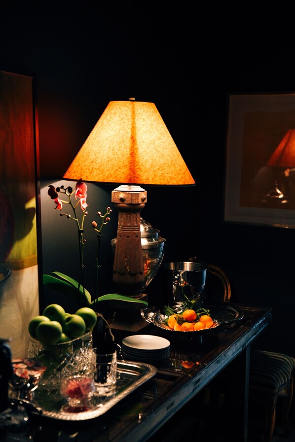 A dimly lit bar area with a table set for a meal, featuring a lamp, a vase with flowers, and a tray with fruits and a drink. - Aida Hotel (Kansas City)