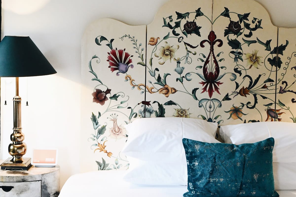 A neatly made bed with white linens and a teal accent pillow, a bedside lamp, and a wall with a floral headboard.