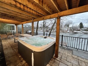 A wooden deck with a hot tub, outdoor furniture, and a view of a river and houses. - Peaceful LakeHouse w/ Spa, Lake Views/Access, and Game Room (Hopatcong)