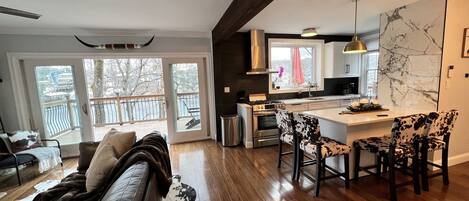A modern kitchen with a central island, a dining area with patterned chairs, and a living room with a sofa and a dog.
