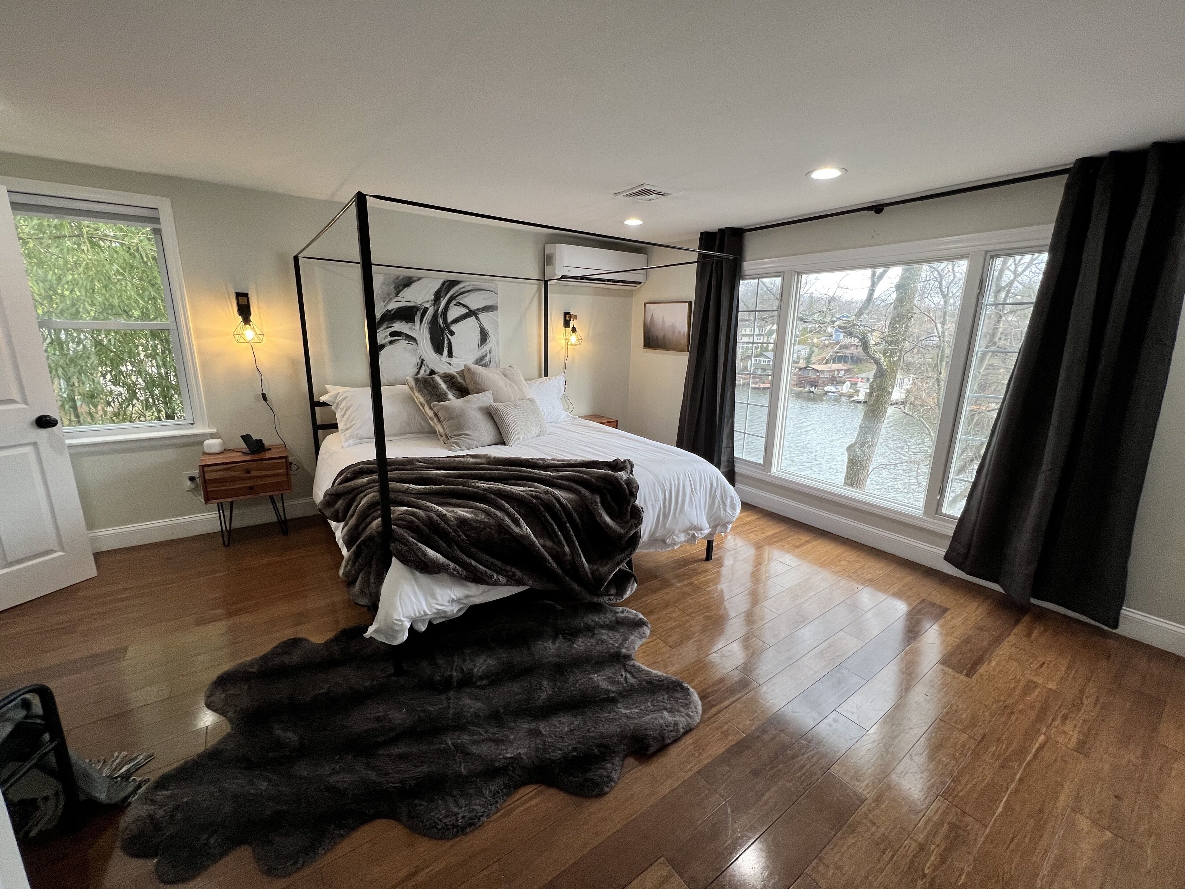 A bedroom with a four-poster bed, a large window with a view of a tree and buildings, and a wooden floor.