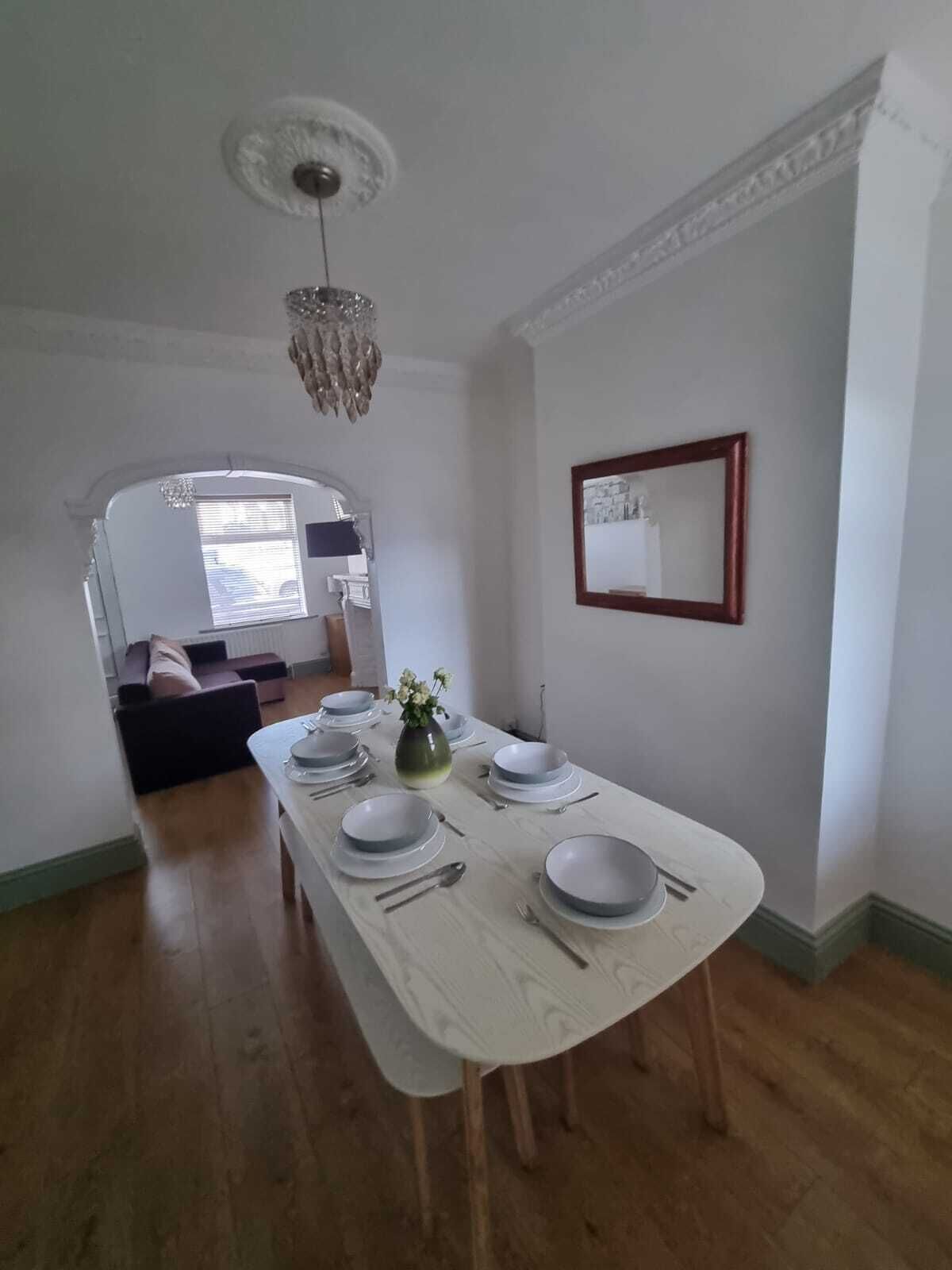 A dining area with a long table set for four, a chandelier, a mirror, and a view into another room.