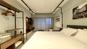 Una habitación de hotel moderna con una cama grande, un banco de madera con espacio de almacenamiento, un televisor en un estante de madera y vistas a la ciudad desde la ventana.
