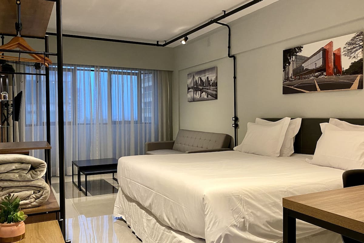 Una habitación de hotel moderna con una cama grande, un escritorio, una silla, un perchero y un ventanal con cortinas traslúcidas.