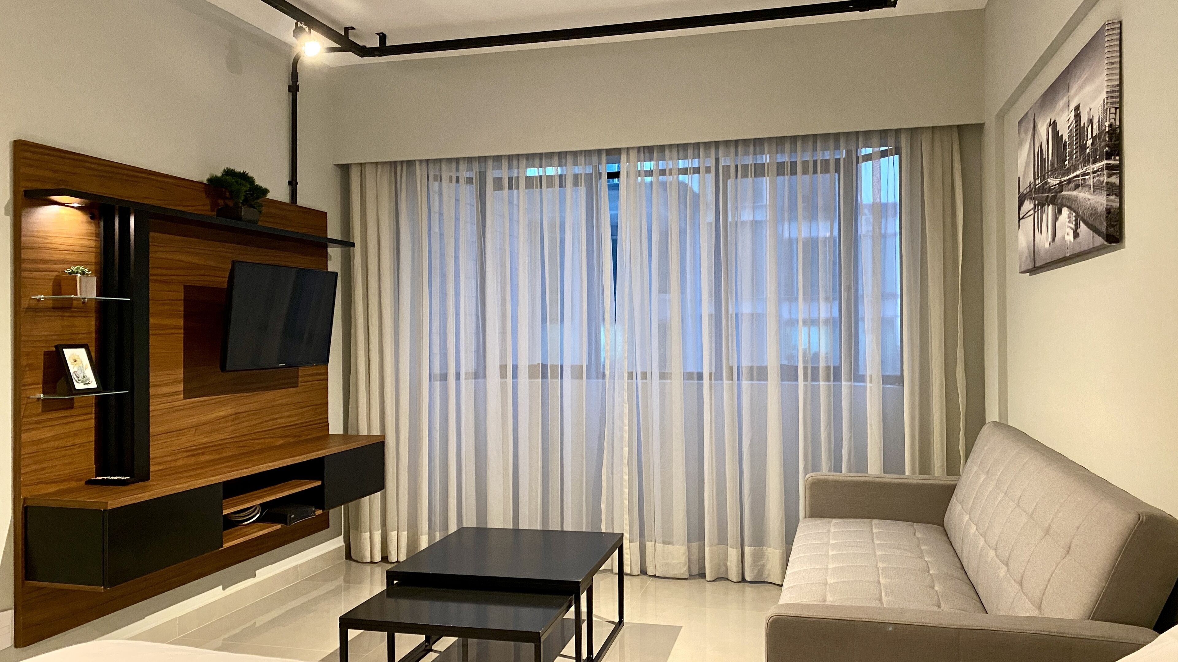 Un soggiorno moderno con un divano, un tavolino, una TV montata su una parete in legno e tende leggere alla finestra.