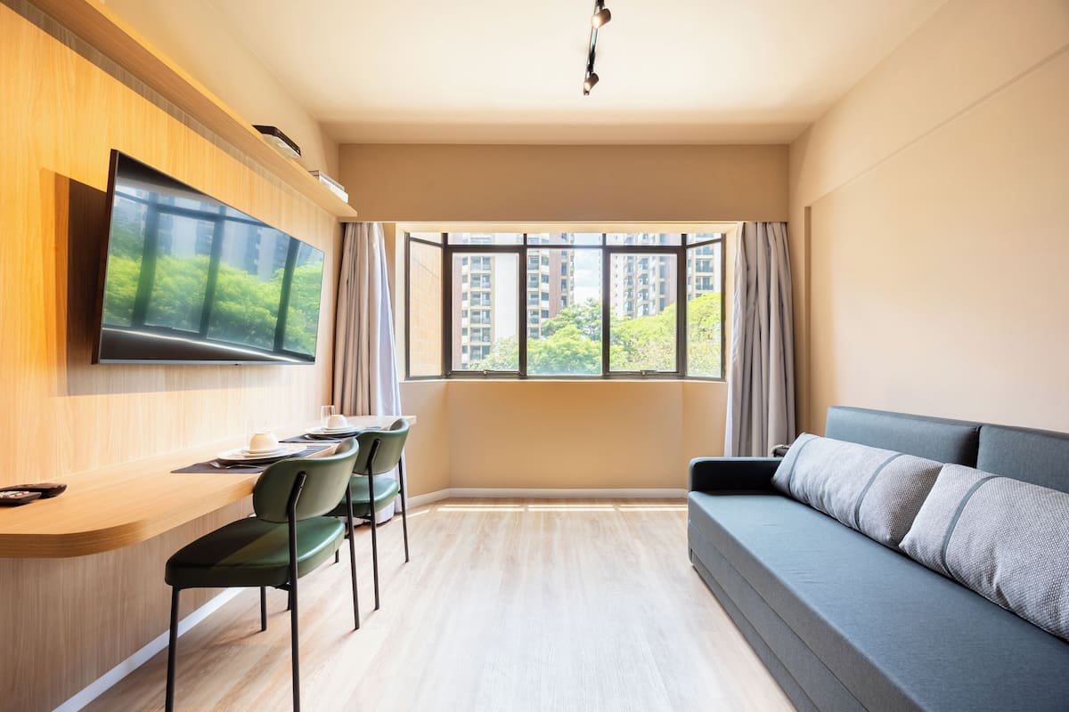 Una sala de estar moderna con un televisor de pantalla plana montado en la pared, un comedor con sillas y una mesa, un sofá con cojines, y un amplio ventanal con vistas a edificios y zonas verdes.