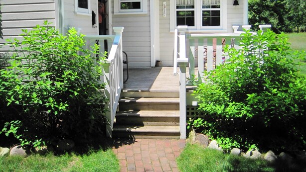 Una casa a due piani con un portico, una scalinata che conduce all'ingresso e un piccolo giardino con cespugli.