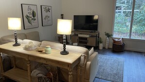 Ein Wohnzimmer mit einem Holztisch, zwei Lampen, einem Fernseher und gerahmten Pflanzenbildern an der Wand.