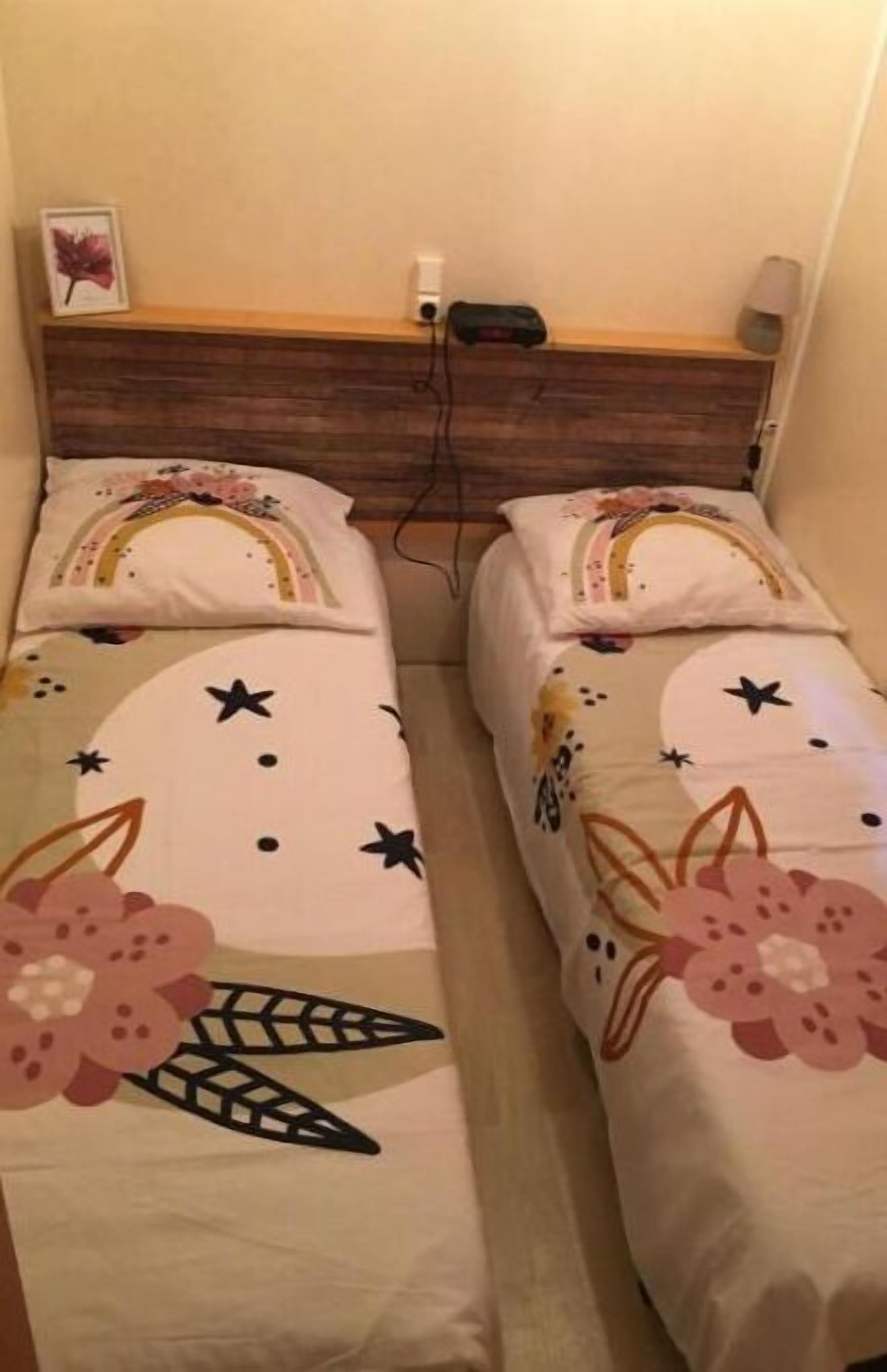 Deux lits jumeaux ornés de motifs floraux et d’étoiles, une tête de lit en bois, une lampe de chevet et un tableau encadré au mur.