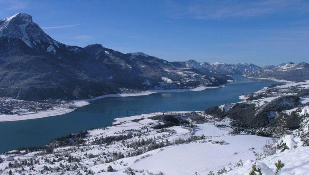 A snow-covered landscape with mountains, a lake, and a bridge.