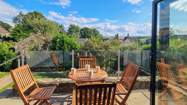 A wooden table and chairs on a patio with a glass railing, overlooking a garden and buildings. - 3 bedroom accommodation in Summerbridge, near Pateley Bridge (Summerbridge, near Pateley Bridge)