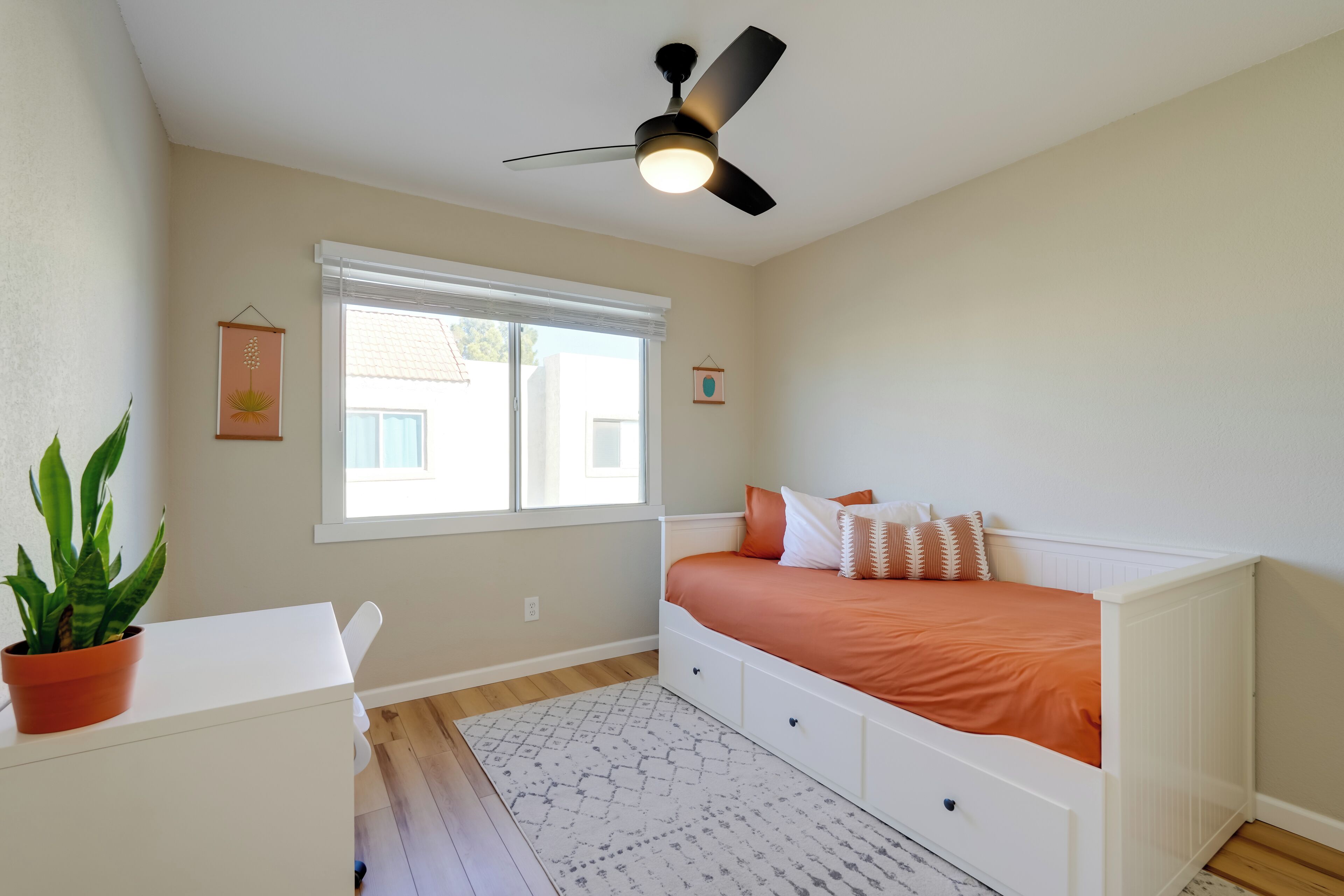 A bedroom with a bed, a ceiling fan, a window, a plant, and a white dresser.