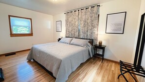 Une chambre à coucher avec un lit, un plancher en bois, une fenêtre, une table de chevet, une lampe et une chaise.