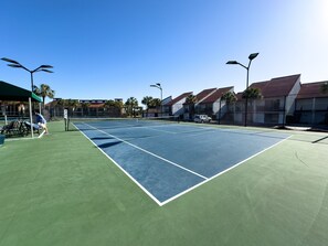 Un campo da tennis all'aperto con una rete, circondato da edifici e palme.