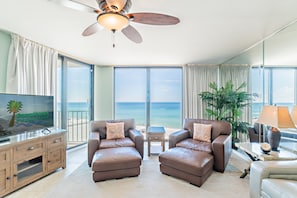 A spacious living room with a large window offering a view of the ocean, a ceiling fan, and a television on a wooden cabinet. - Solace-Edgewater 613T3 Beachfront-Fully Renovated! Luxurious Walk-In Shower! (Panama City Beach)