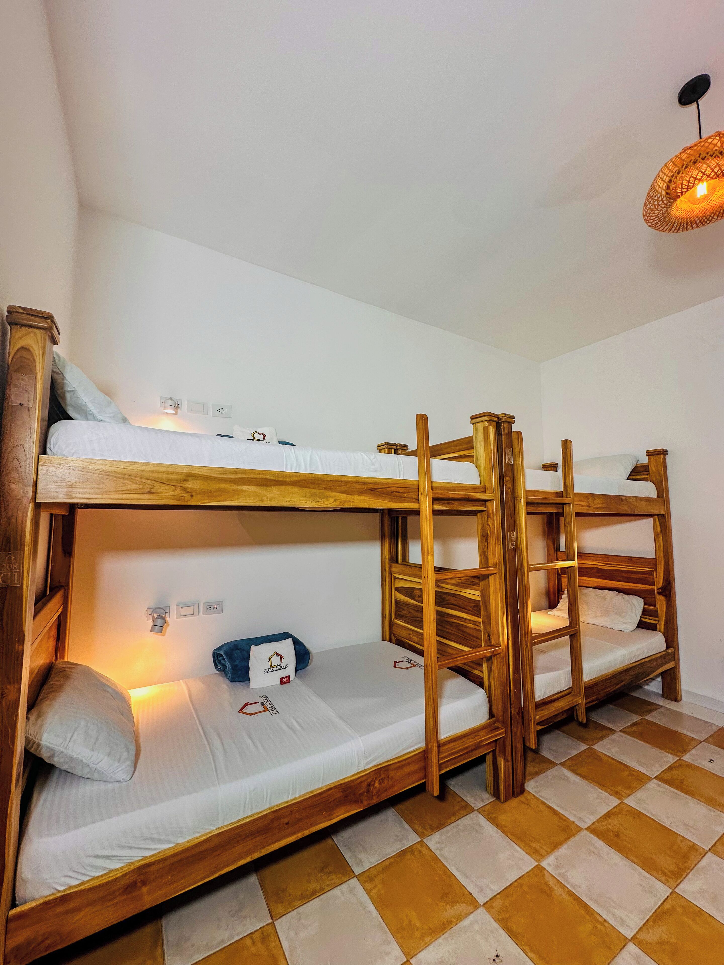 Une chambre avec des lits superposés, des structures en bois, une literie blanche et un sol à carreaux.