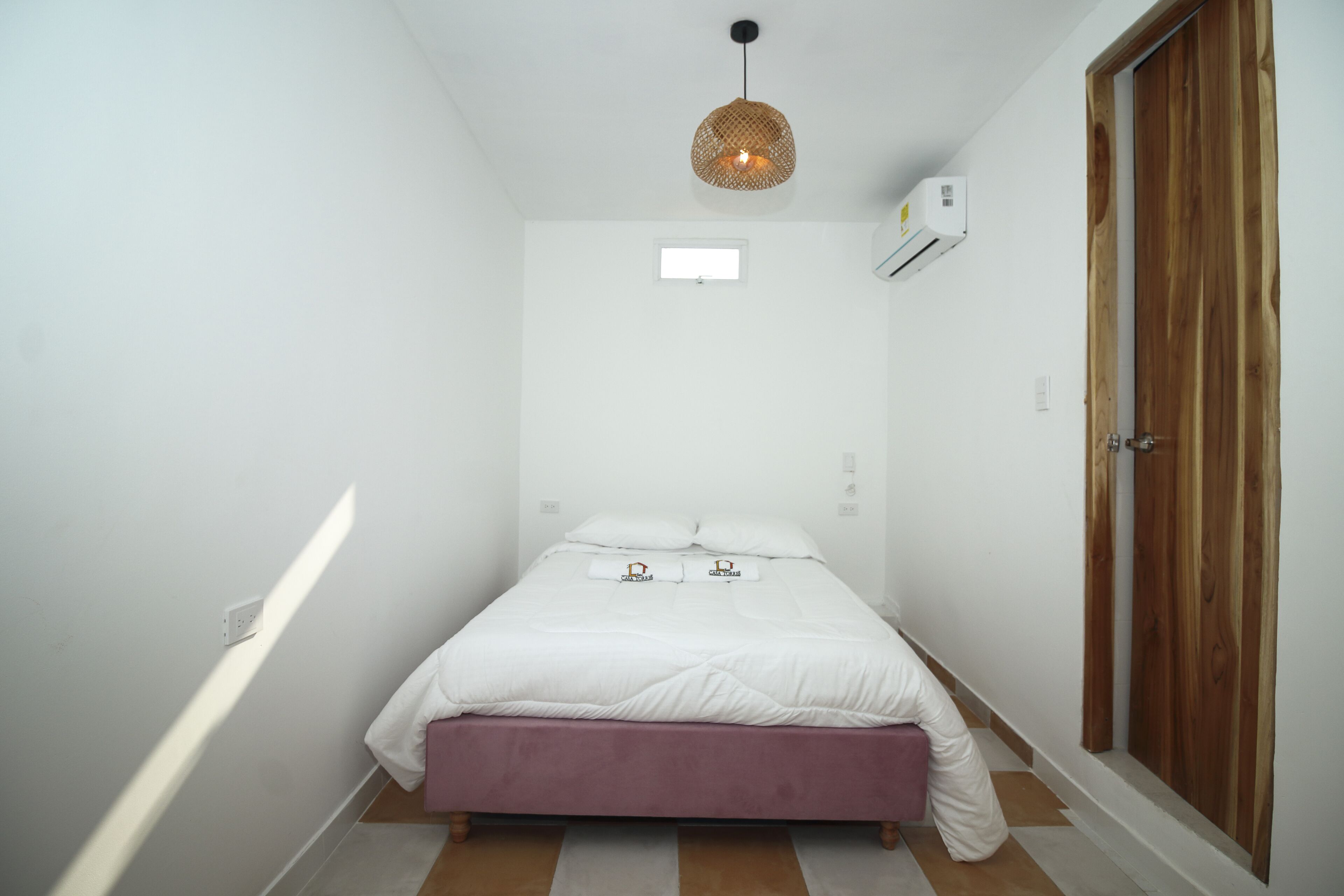 A small, neatly arranged bedroom with a single bed, a wooden wardrobe, a hanging light fixture, and a small window.
