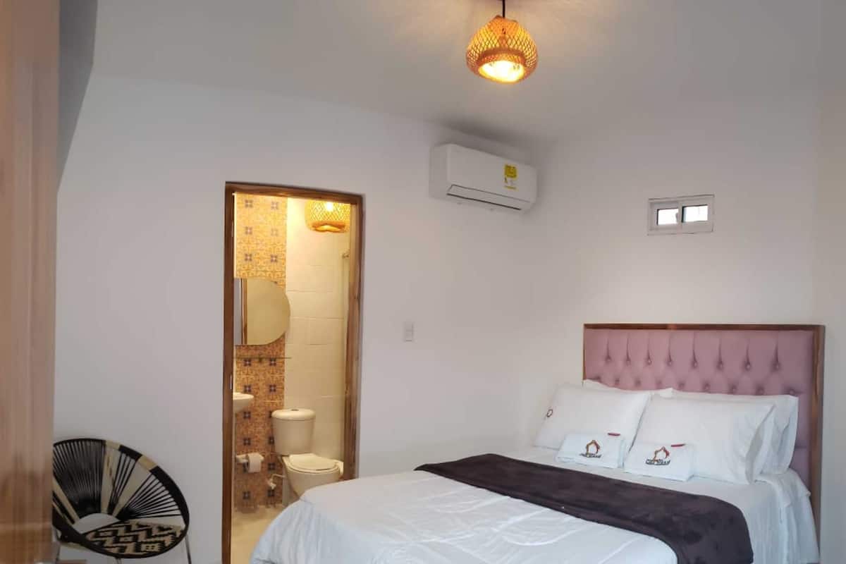 A bedroom with a bed, a chair, a bathroom visible through an open door, and an air conditioning unit on the wall.