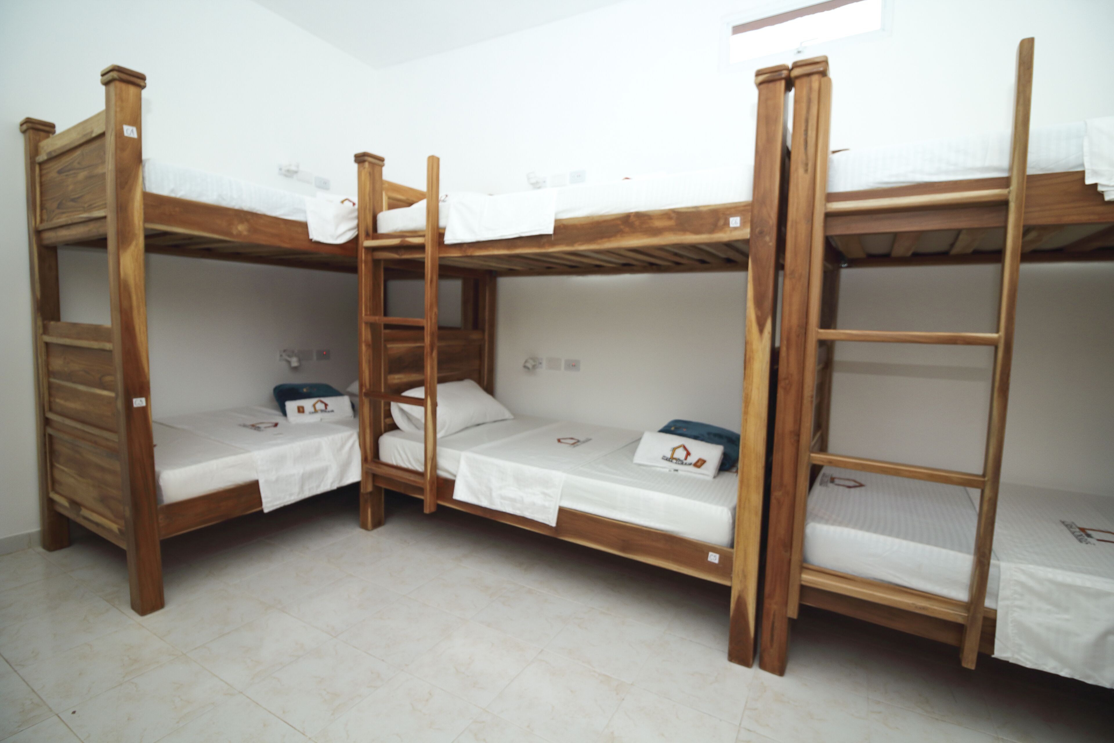 A room with four wooden bunk beds, each with a white mattress and pillow. There is a window providing natural light, and the floor is tiled.