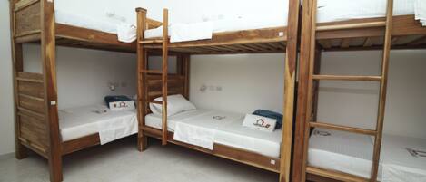 A room with four wooden bunk beds, each with a white mattress and pillow. There is a window providing natural light, and the floor is tiled.