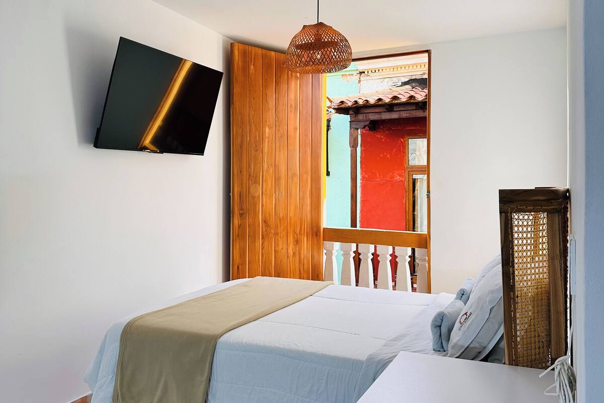 A bedroom with a bed, a bedside table, a TV mounted on the wall, and a window with a view of a building.