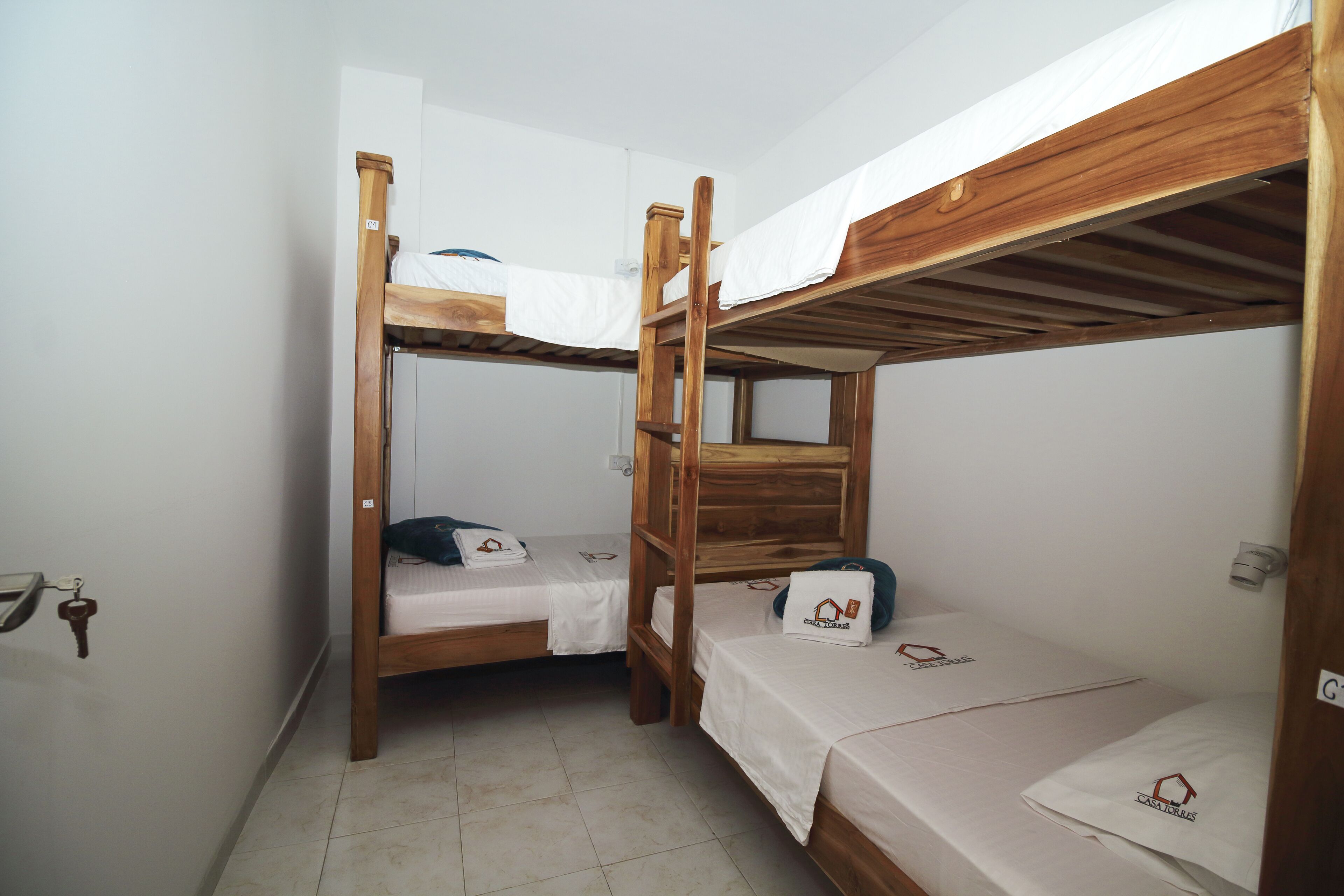 Una habitación con dos literas, cada una con estructura de madera y ropa de cama blanca. Hay una pequeña mesa con una silla frente a una de las camas, y un estante montado en la pared sobre la otra cama.