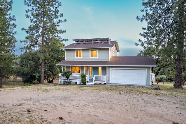 A two-story house with a garage, surrounded by trees and a dirt driveway.