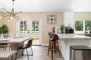 A modern kitchen with a large island, bar stools, a dining area with a table and chairs, and a view through the glass doors.
