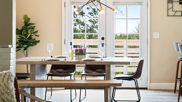 Uma sala de jantar com mesa de madeira, duas cadeiras, um candelabro e vista através de uma porta de vidro.
