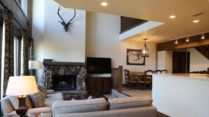 A living room with a stone fireplace, a mounted deer head, a television, and a dining area with a chandelier. - Viking Two Bedroom Loft 2 Apartment Hotel by RedAwning (Park City)