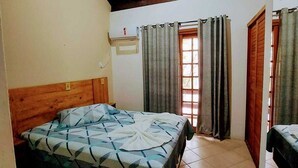 Un dormitorio con cabecera de madera, cama con colcha de estampado geométrico, un estante con objetos, una ventana con cortinas y una puerta.