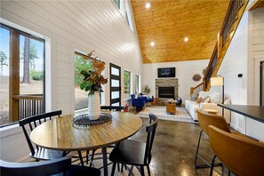 Amplia sala de estar con techo de madera, chimenea y comedor con mesa y sillas redondas.