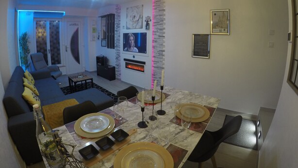 A modern living room with a sofa, TV, and a dining area with a table set for two. - hébergement au bercail (Beaumont-sur-Sarthe)