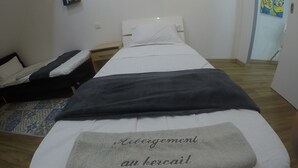A single bed with a grey blanket and a towel with text, a bedside table, a nightstand, and a wall with a picture. - hébergement au bercail (Beaumont-sur-Sarthe)