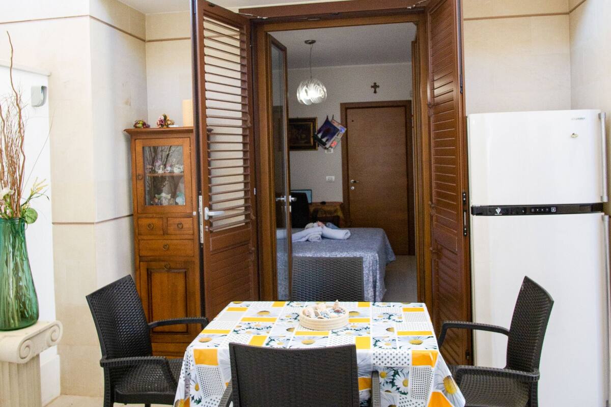 A dining area with a table set for two, two chairs, a cabinet, and a refrigerator.